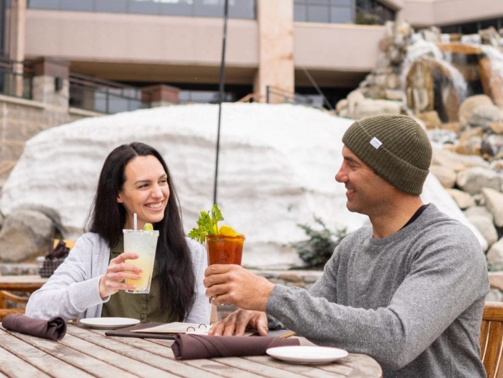 Man in wool hat and woman cheers cocktails sitting outside at Everline Resort and Spa restaurant during winter