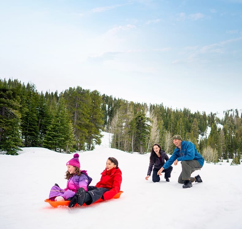 Mother and father laugh as they push two kids on a plastic sled down a snowy hill in Lake Tahoe