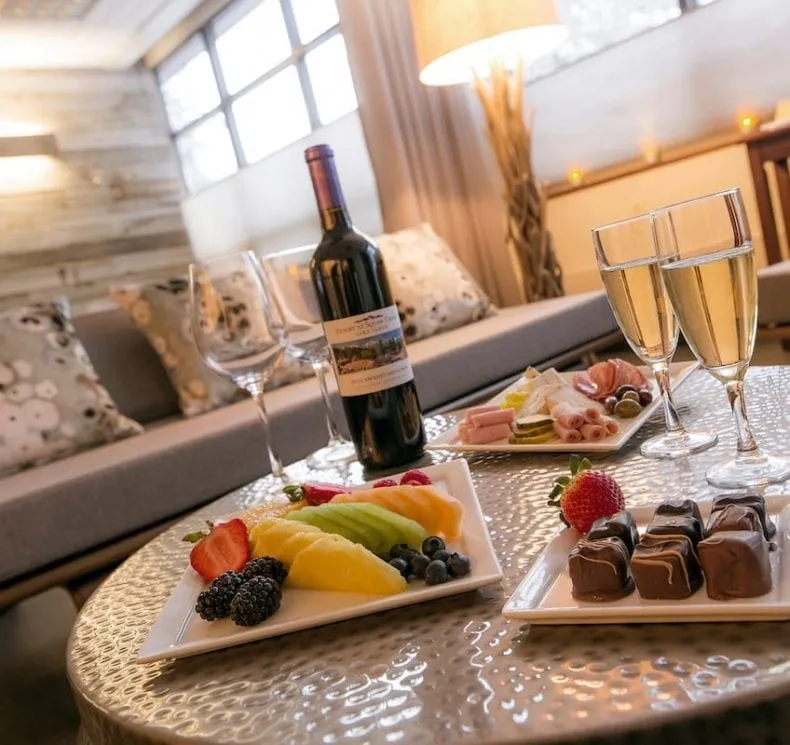 A fruit platter, chocolates and charcuterie are beautifully plated beside a bottle of wine and several glasses.