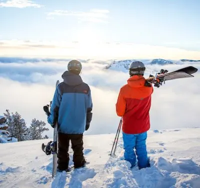 Two people holding their snowboard and skis look down on clouds from atop Lake Tahoe mountain peak